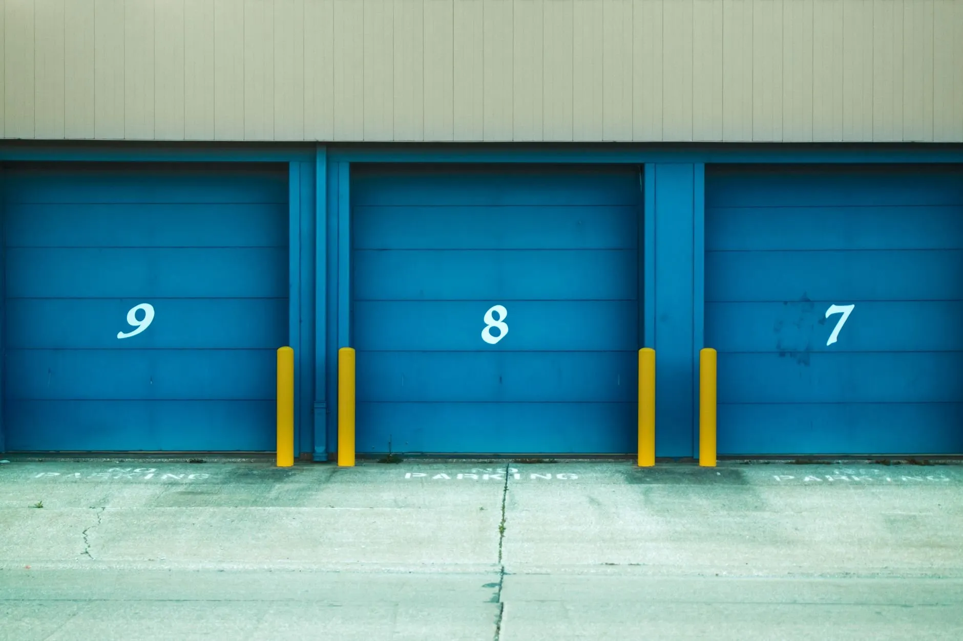 Three blue roller doors numbered 9, 8 and 7 with yellow bollards and a painted no-parking line on the concrete in front