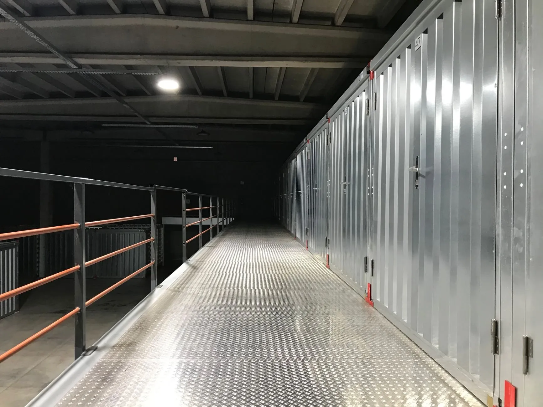 A lit chequer-plate walkway on the mezzanine running past a long row of steel unit doors