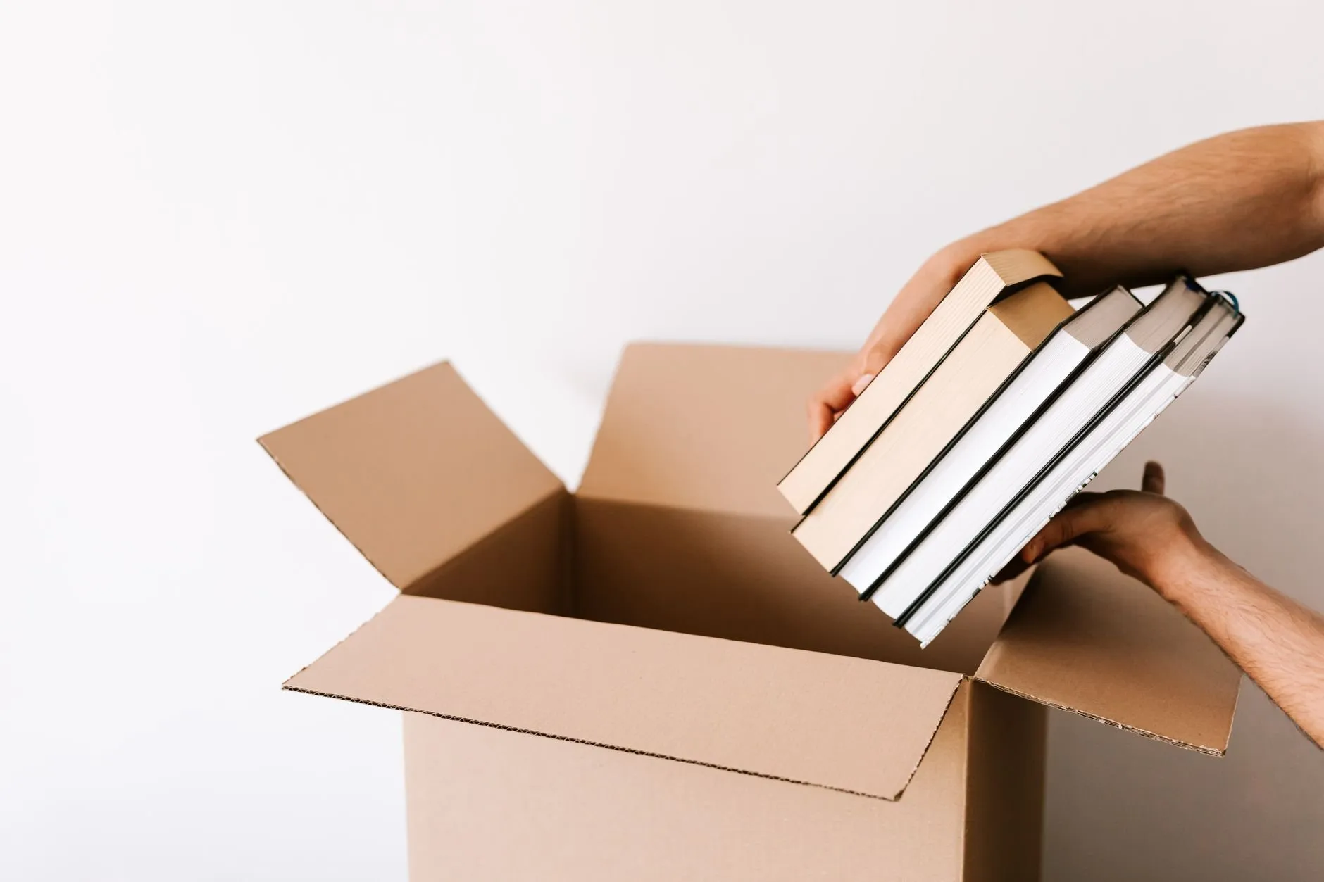 Hands lowering a stack of hardback books into an open cardboard box against a white wall