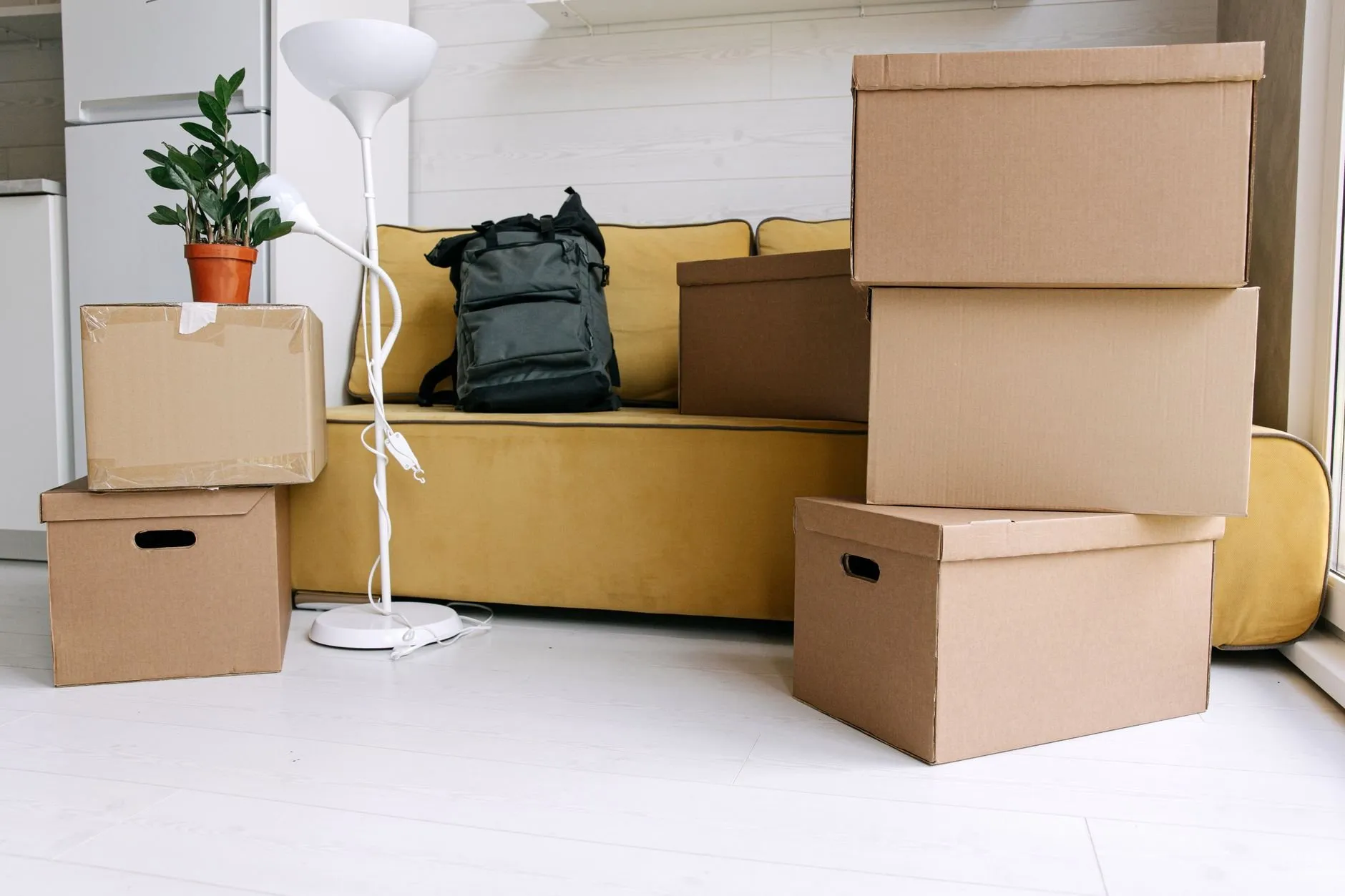 A mustard sofa in a bright room with cardboard archive boxes stacked beside it and a floor lamp waiting to be packed